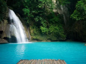 A close-up view of a tranquil waterfall cascading into a pool of blue water, framed by dense tropical forest.