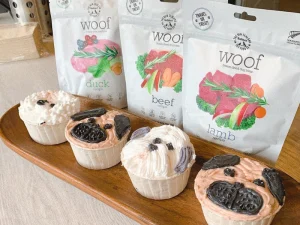 A wooden tray displaying decorated pet-friendly cupcakes shaped like dogs, with bags of freeze-dried dog treats in the background.