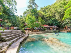 A vibrant scene of people enjoying a natural spring with turquoise water, surrounded by trees and small structures for relaxation.