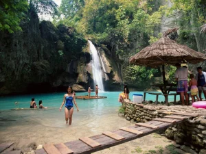 Tourists swimming and relaxing near a picturesque waterfall, with a wooden gazebo and rocky pathways nearby.