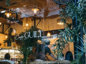 The exterior of Café Guilt, featuring large glass windows and green plants surrounding the cafe, with people dining inside.
