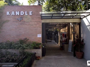 The entrance to Kandle Café, showcasing a modern brick facade with greenery and a welcoming pathway leading inside.