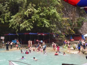 A lively gathering of visitors in a spring with shallow turquoise water, colorful umbrellas, and trees providing shade