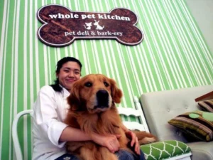 A woman sitting with a golden retriever on a couch inside Whole Pet Kitchen, a pet-friendly deli and barkery, with the cafe logo displayed on the wall.