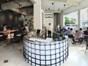 The interior of Commune Café, featuring an industrial design with a curved bar counter, large windows, and customers working or chatting at tables