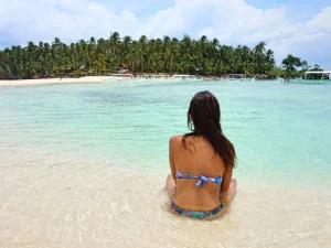 A woman in a colorful bikini sits on the shoreline, gazing at the clear blue waters and a tropical island covered with coconut trees in the background