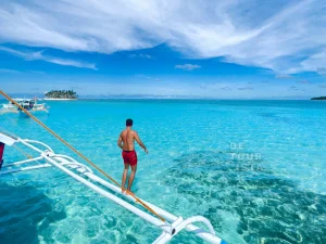A man in red swim shorts stands at the edge of a boat, ready to jump into the transparent, shallow ocean with an island in the background