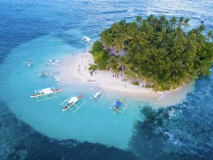 Aerial view of a small white sand island covered in greenery, surrounded by boats anchored in vibrant blue waters with coral reefs visible below