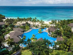 MovenPick Resort - Travelpreneur A tropical beachfront resort with a large, lagoon-style pool surrounded by lush greenery, palm trees, and direct access to a white sand beach with turquoise waters.