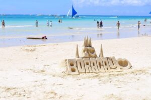 boracay island - Travelpreneur A sandcastle with the word "BORACAY" carved into it on a white sandy beach, with people, sailboats, and clear blue waters in the background.