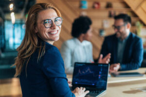 a woman in professioanl clother smilling at the camera while her laptop shows that she is doing forex trading