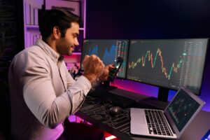 a man doing a happy gesture with his fist closed and arms raised as if saying he did it in front of a monitor presenting forex trading data