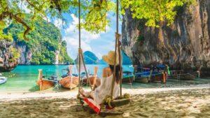 thailand - Travelpreneur a woman sitting on a swing looking facing the beautiful beach with boats near the shore