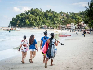 Group of friends walk along tropical beach lined with boats and resorts, enjoying island tourism