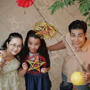 Family smiles holding parol lanterns indoors, festive decor above, Holidays in the Philippines celebration together