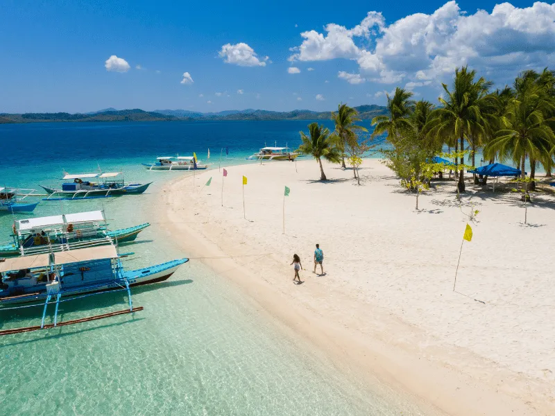 Couple walks sandbar beside outrigger boats and palms, Holidays in the Philippines island getaway view