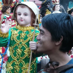 Man smiles beside Santo Niño statue, Holidays in the Philippines devotion display among festival figures