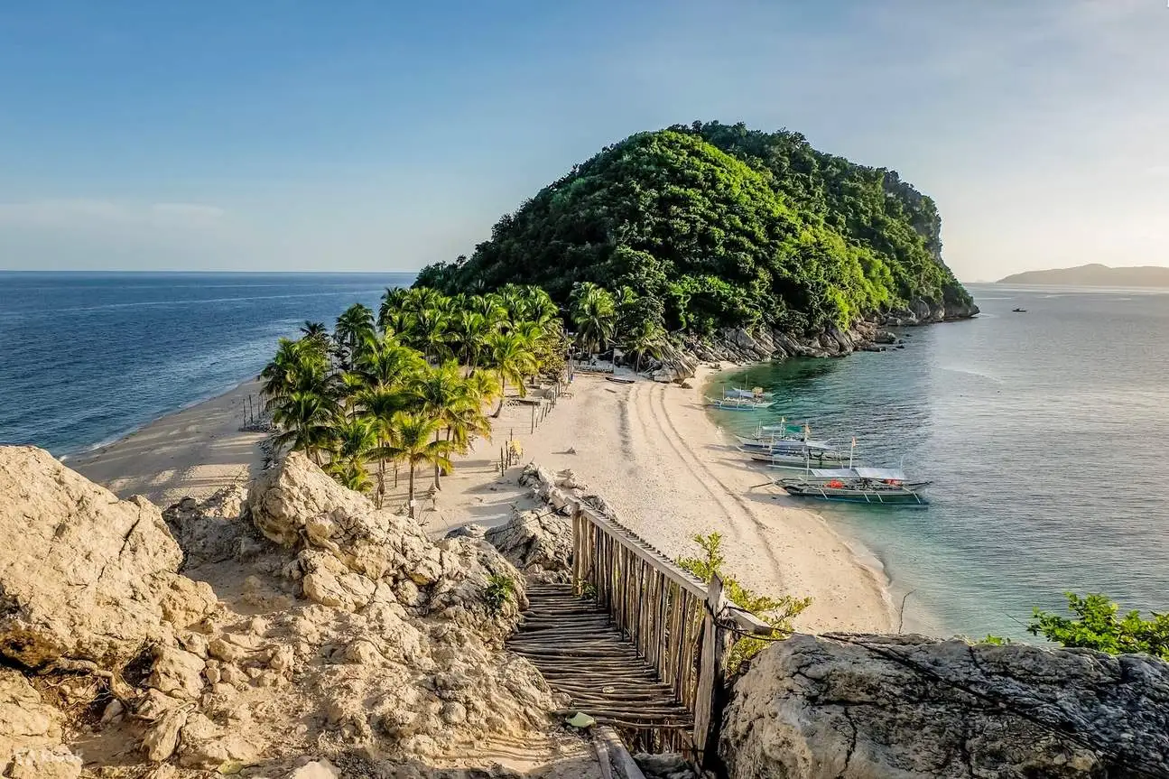Cabugao Gamay Island in Islas de Gigantes, Iloilo - Travelpreneur High-angle view of Cabugao Gamay Island in Gigantes, showing a sandy beach strip lined with coconut palms, outrigger boats, and a lush green hill under a clear blue sky.