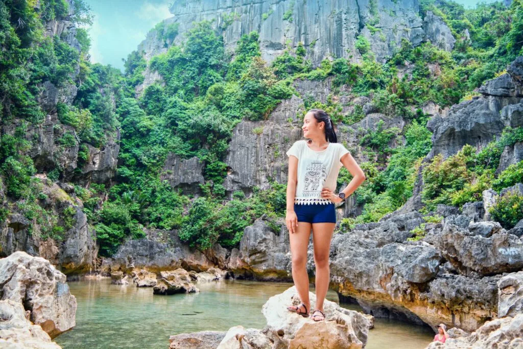 Inside Tangke Saltwater Lagoon, Islas de Gigantes - Travelpreneur A female traveler posing on a rocky outcrop inside Tangke Saltwater Lagoon, surrounded by towering, lush green limestone cliffs and a shallow natural pool.
