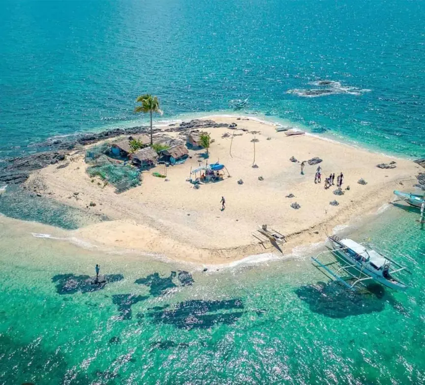 Aerial View of Pulupandan Island, Carles, Iloilo - Travelpreneur Drone shot of Pulupandan Island in Gigantes, a tiny white-sand islet with a single palm tree, small native huts, and motorized outrigger boats anchored in clear turquoise water.