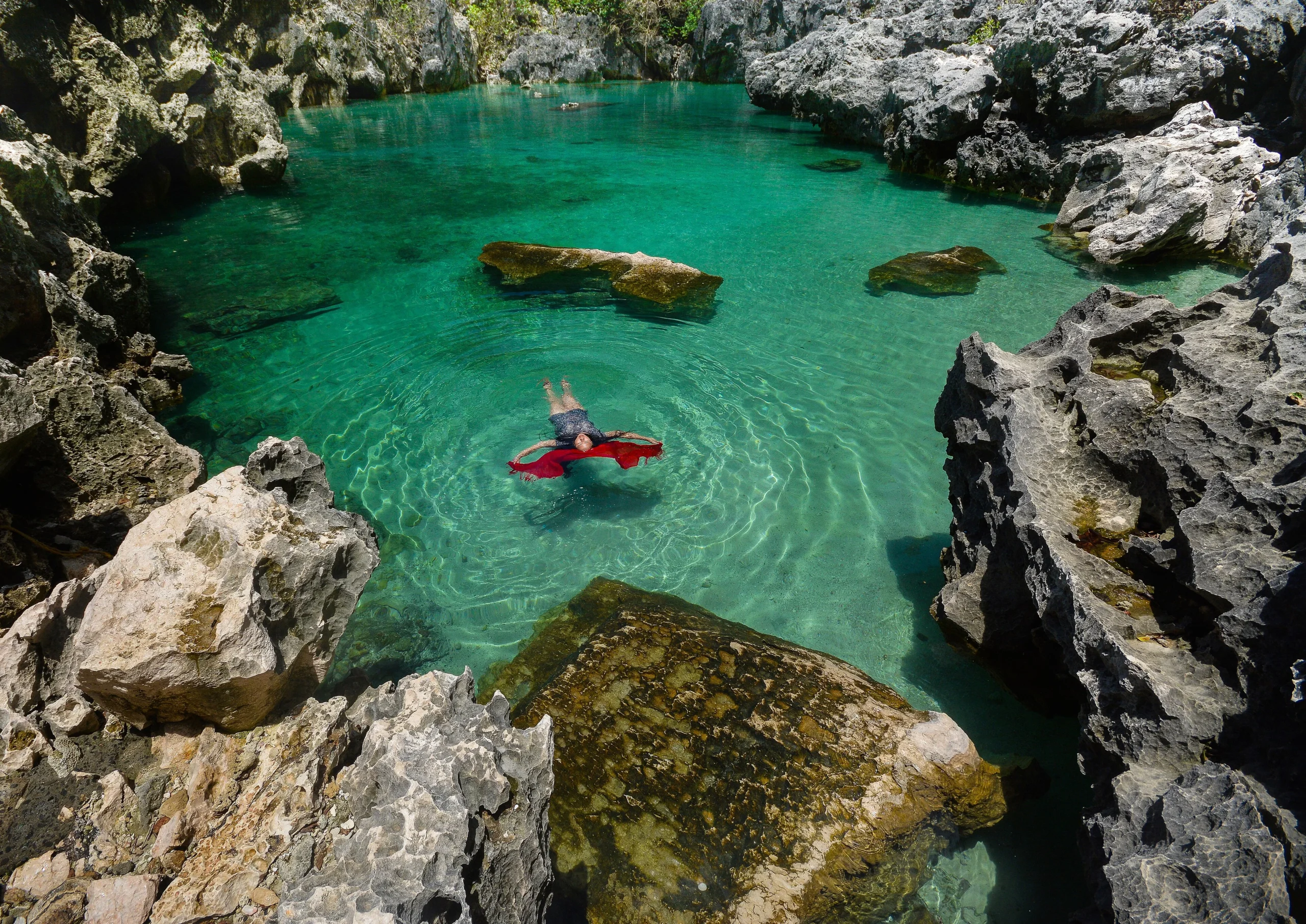 Tangke Saltwater Lagoon in Islas de Gigantes, Iloilo - Travelpreneur A traveler in a swim suit floating on her back with a red sarong in the crystal-clear emerald waters of Tangke Saltwater Lagoon, surrounded by steep jagged limestone cliffs.