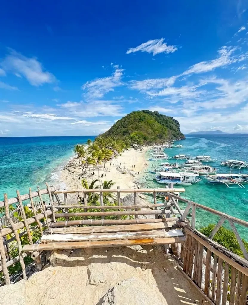 Panoramic view of Cabugao Gamay Island from a wooden viewing deck, showing a narrow white sand beach lined with palm trees and outrigger boats in turquoise water.