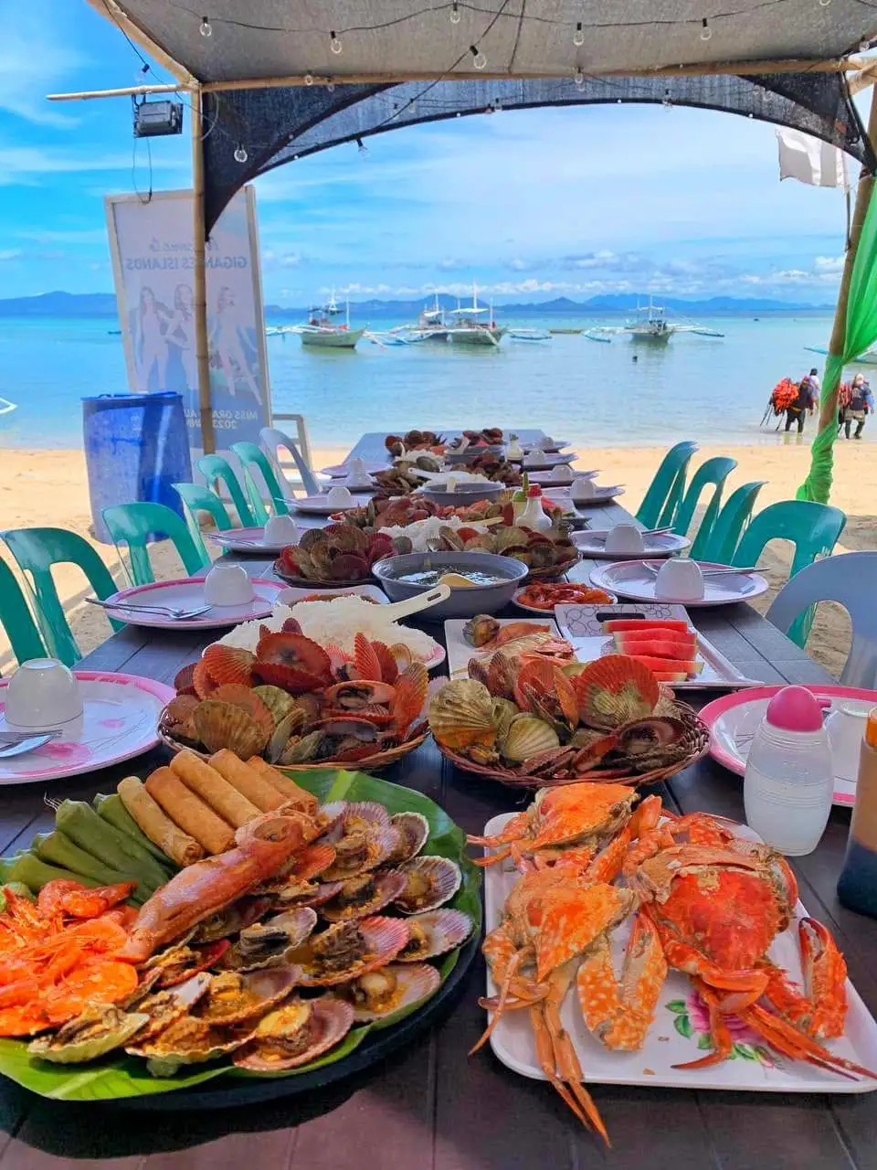 A long beachfront dining table filled with a Filipino seafood feast including fresh crabs, steamed scallops, shrimp, and lumpia, with the ocean and outrigger boats in the background.