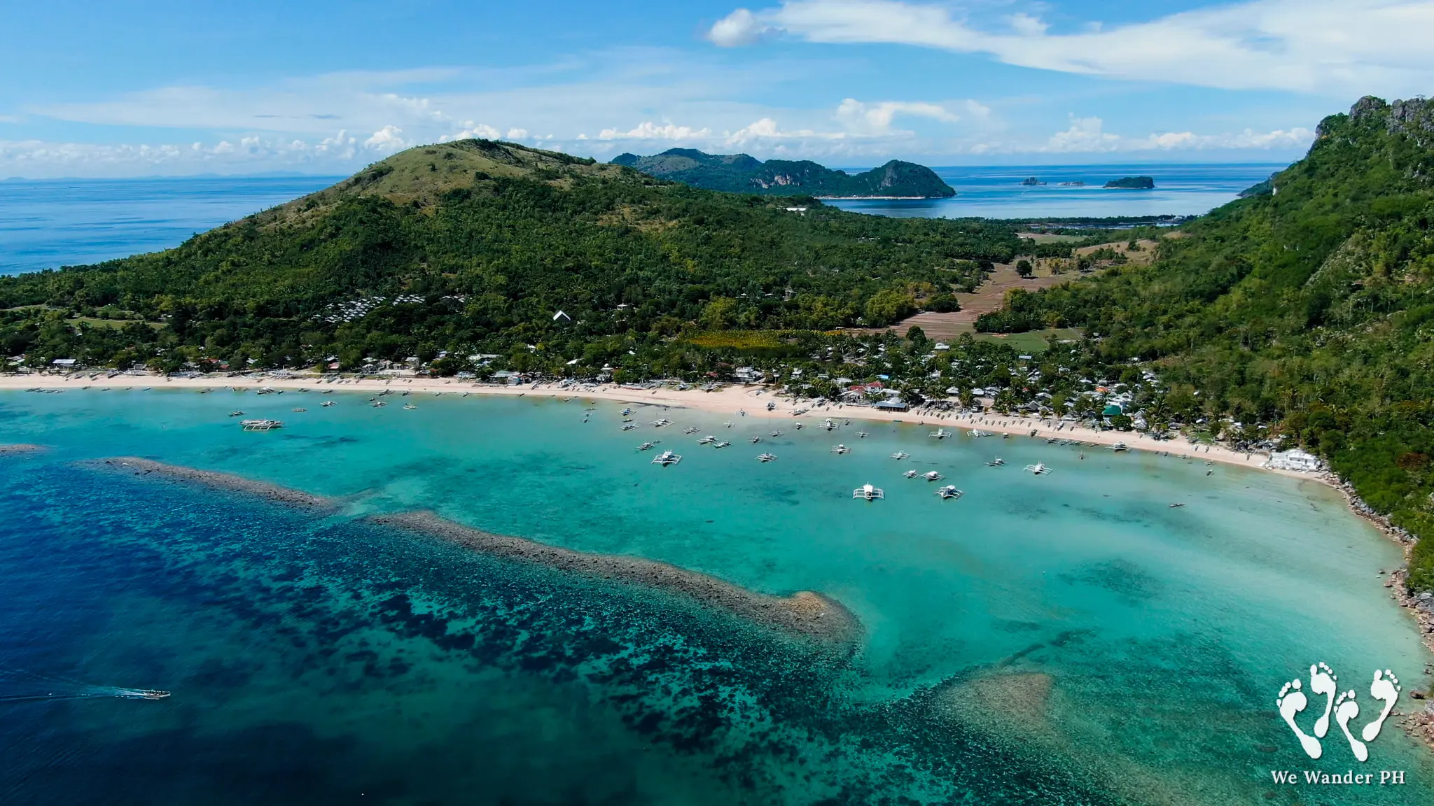 Lantangan Beach, Gigantes Sur, Iloilo - Travelpreneur Panoramic drone shot of Lantangan Beach on Gigantes Sur Island, showcasing a long sandy shoreline lined with coastal houses, a fleet of small outrigger fishing boats, clear turquoise waters, a vibrant coral reef, and rolling green hills under a blue sky.
